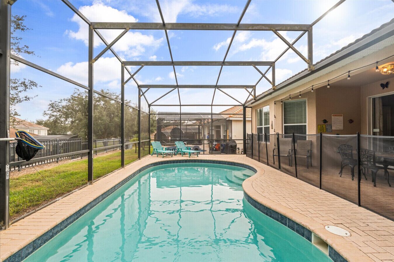 A covered outdoor swimming pool with a pergola structure, surrounded by a fence, and a patio area with chairs and tables.
