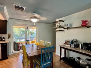 A kitchen with a wooden table, chairs, and a ceiling fan. There are shelves with kitchen items and a window with a view of greenery.