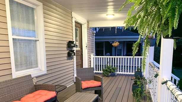 Une terrasse extérieure couverte, aménagée avec des meubles en osier, des plantes en pot et une rambarde blanche.