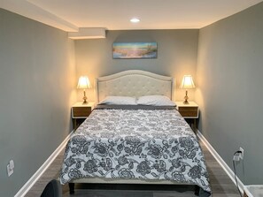 A bedroom with a bed, two bedside tables, a nightstand, a wall-mounted painting, and a power outlet. - Semi Private cozy studio near downtown. (Montclair)