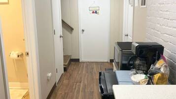 A narrow hallway with wooden flooring, a white door with a hanging coat rack, and a kitchenette area with a coffee maker and a microwave.
