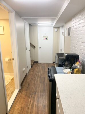 Un pasillo estrecho con suelo de madera, una puerta blanca con perchero y una pequeña cocina con cafetera y microondas.