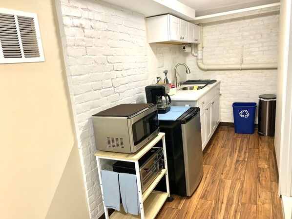 Una cocina compacta con paredes de ladrillo blanco, suelo de madera y electrodomésticos básicos como microondas, tostadora y refrigerador.