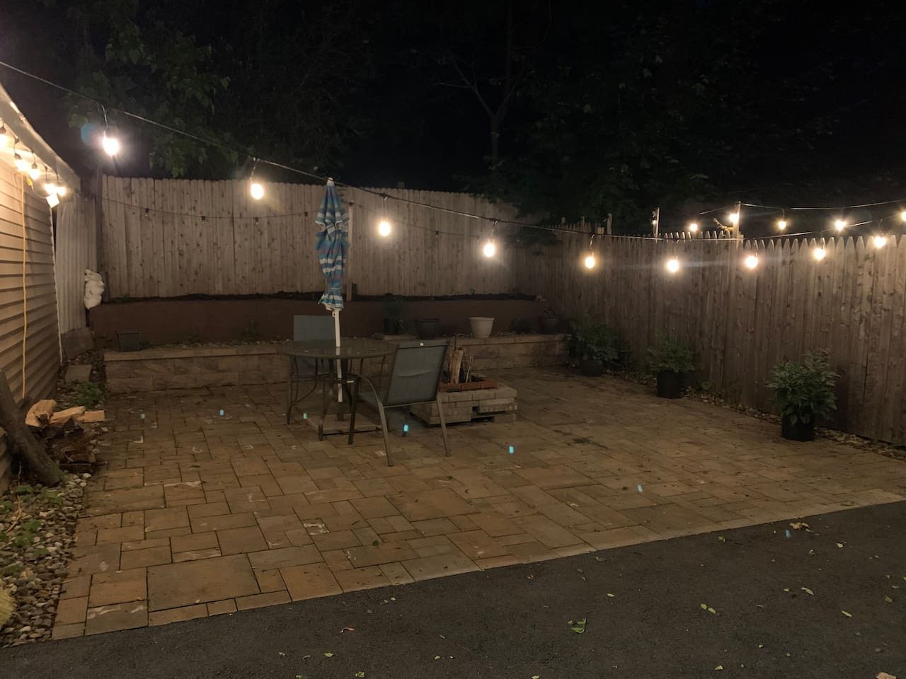 Een achtertuin terras met een metselstenen vloer, een tafel, stoelen en lichtslingers.