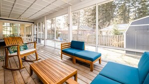 Une terrasse extérieure couverte, aménagée avec des meubles en bois, dont un fauteuil à bascule et un canapé, et de grandes fenêtres offrant une vue sur le jardin arrière.