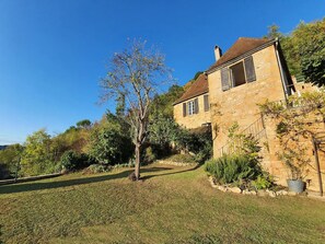 A stone house with a sloped roof, surrounded by a well-maintained garden and a clear blue sky.