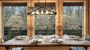 Une table à manger en bois, dressée avec des assiettes, des verres et des serviettes, et offrant une vue sur une colline boisée à travers de grandes fenêtres.