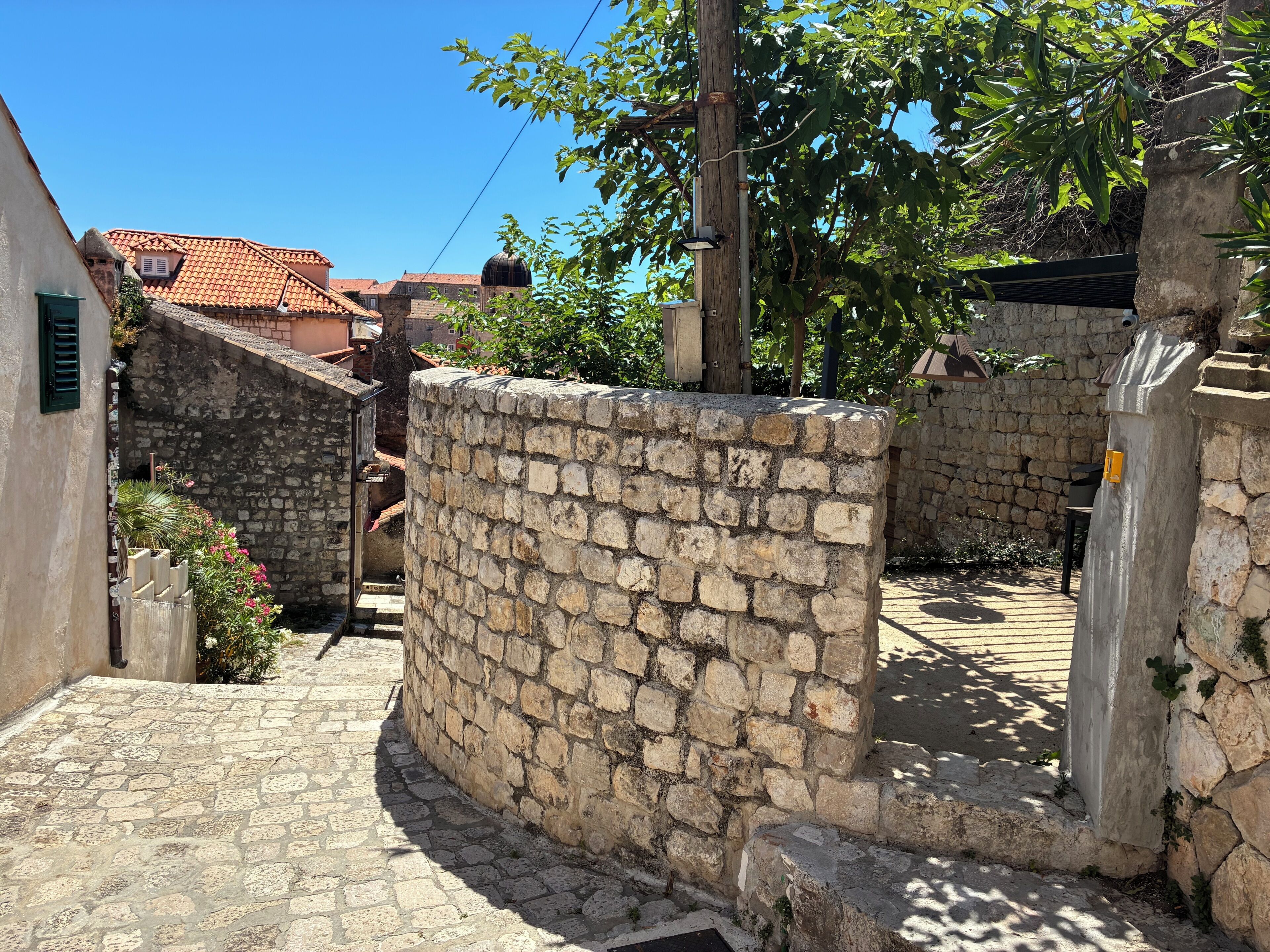 Eine Mauer aus Stein mit gewölbter Oberseite, ein gepflasterter Weg und im Hintergrund ein Gebäude mit einem Ziegeldach.