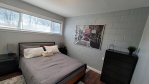 A bedroom with a bed, a nightstand, a dresser, a window with blinds, a wall-mounted picture, and a ceiling light. - Newly remodeled 1 Bedroom apartment (Ypsilanti)