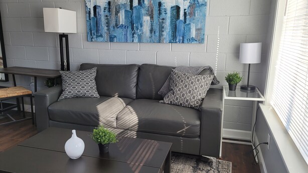 A modern living room with a grey sofa, patterned cushions, a cityscape painting, a small table with a vase, and a floor lamp. - Newly remodeled 1 Bedroom apartment (Ypsilanti)