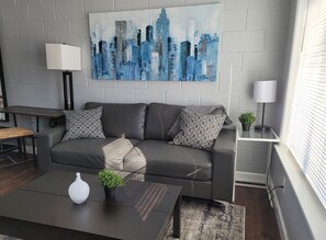 Een moderne woonkamer met een grijze bank, gekleurde kussens, een schilderij van een stadsgezicht, een kleine tafel met een vaas, en een vloerlamp.