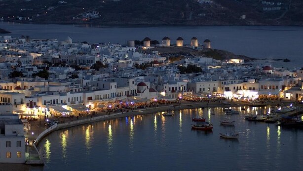 Un pueblo costero con edificios blancos, un puerto con barcos y una colina con construcciones al fondo.