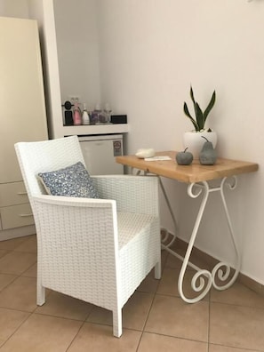 Ein weißer Korbstuhl mit gemustertem Kissen, ein kleiner Holztisch mit einer Pflanze und eine weiße Kommode in einem Raum mit Fliesenboden.