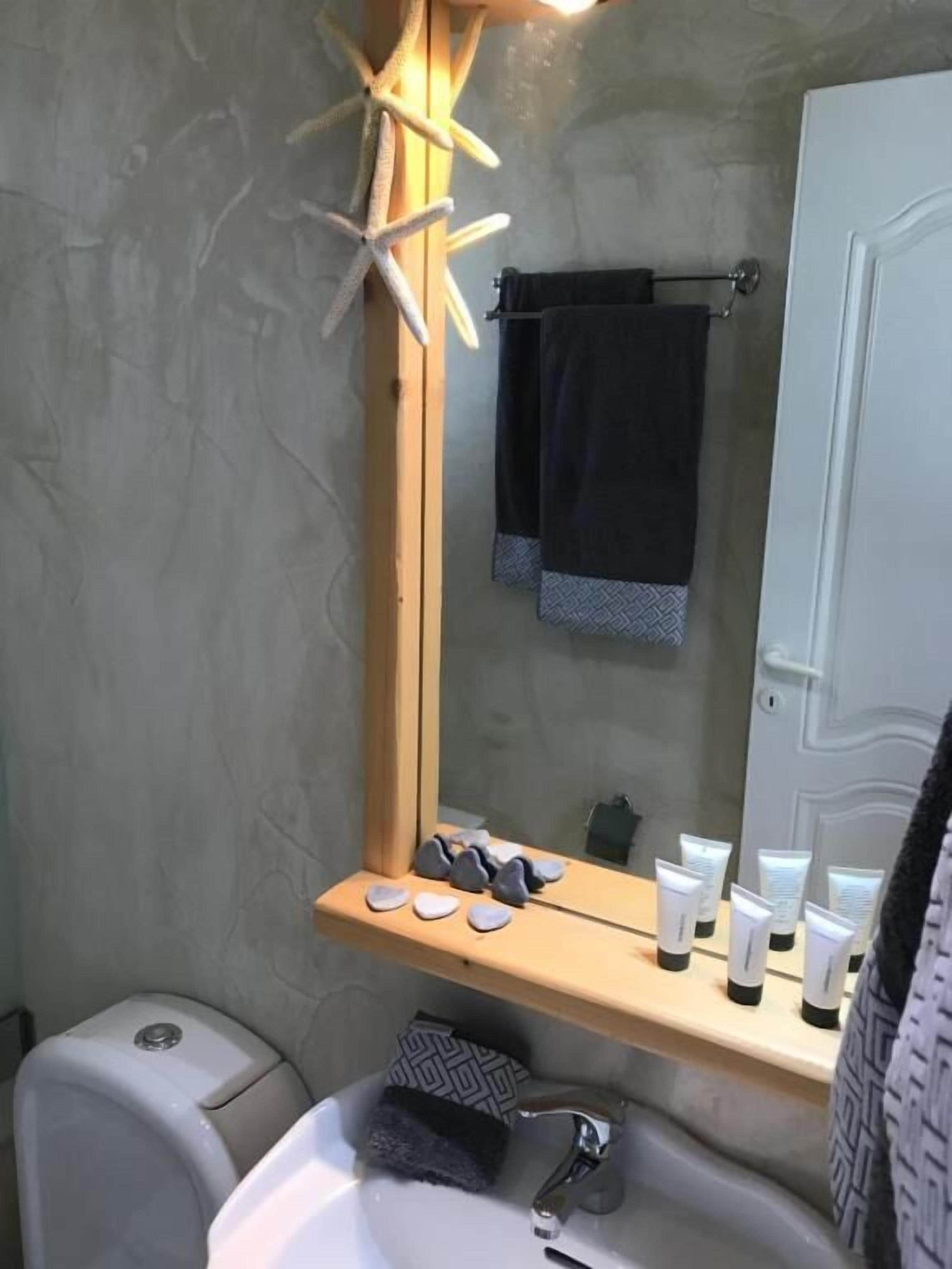 Un baño con lavamanos blanco, espejo, toallero y artículos de aseo en un estante.