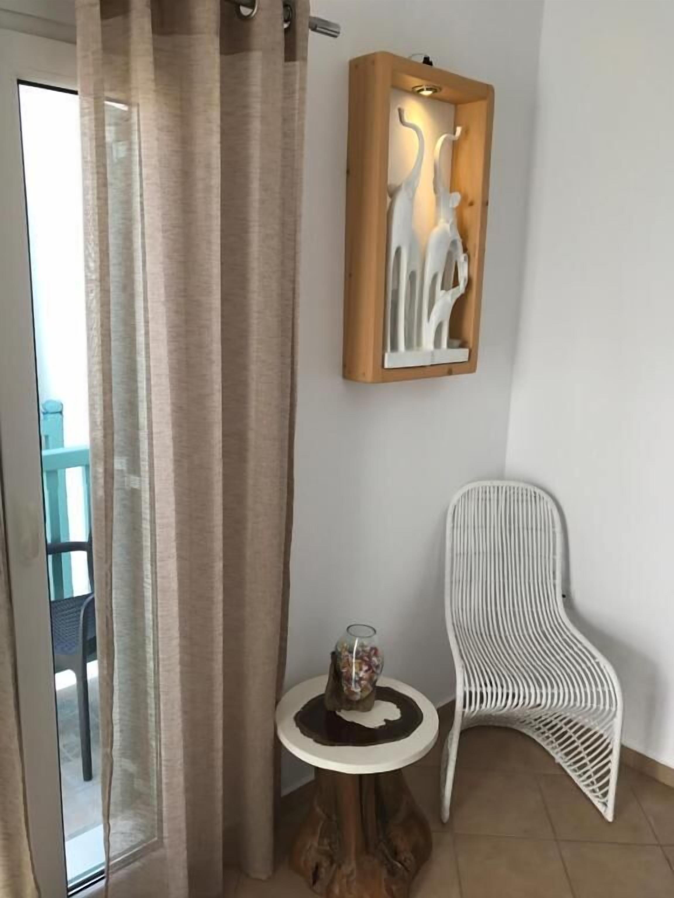 A room with a glass door, a wooden-framed artwork, a white chair, and a small table with a glass object.