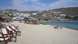 Una playa con hileras de tumbonas de madera y algunas personas tomando el sol. Al fondo, edificios blancos en una colina.