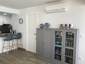 A modern kitchen with a built-in stove, two bar stools, a cabinet with glass doors displaying dishes and glasses, and a wall-mounted air conditioner.