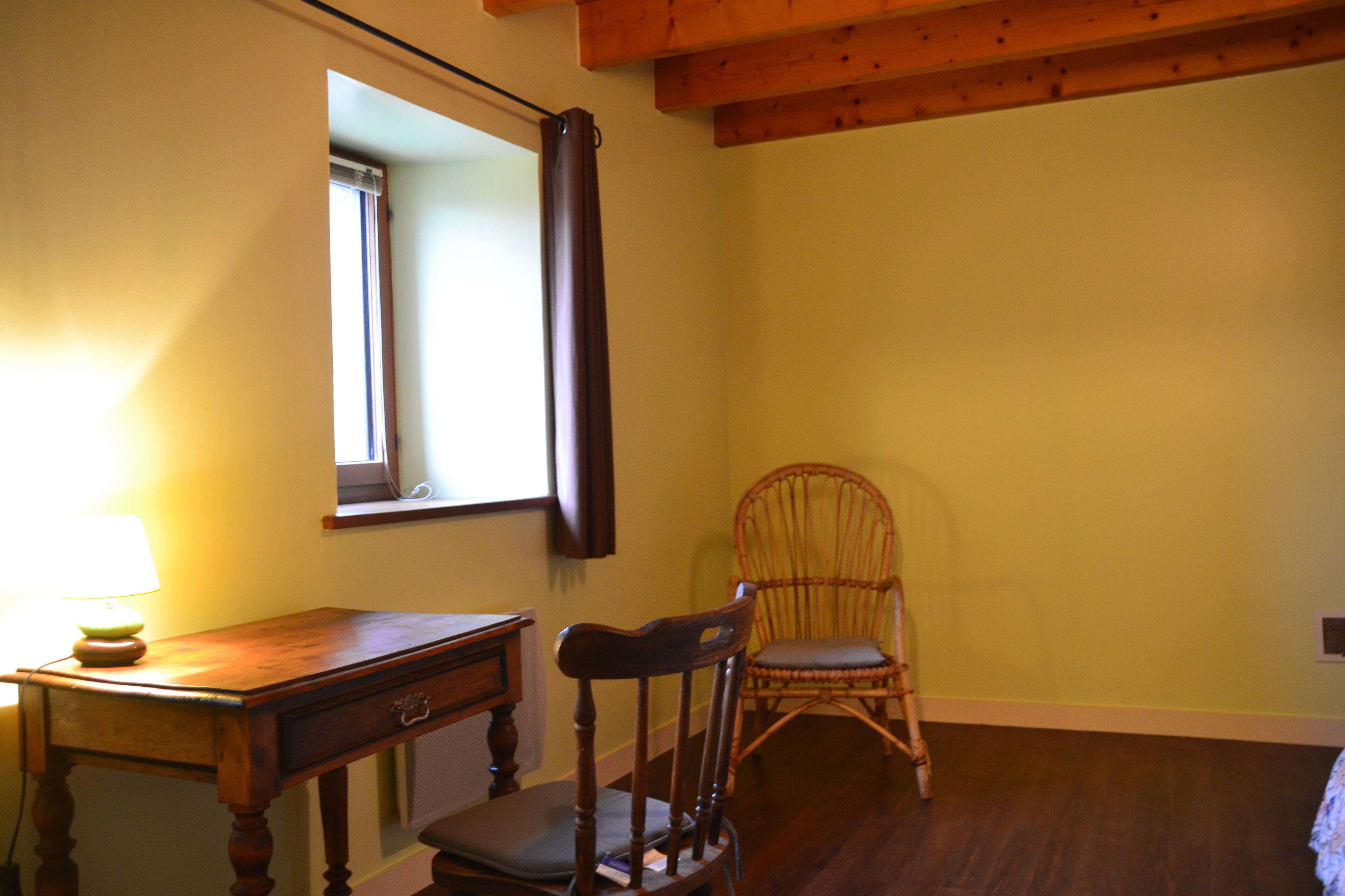 Habitació amb escriptori de fusta, una cadira, finestra amb cortina i biga de sostre de fusta.