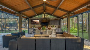 Un patio exterior techado con chimenea de piedra, mobiliario de mimbre y un televisor.