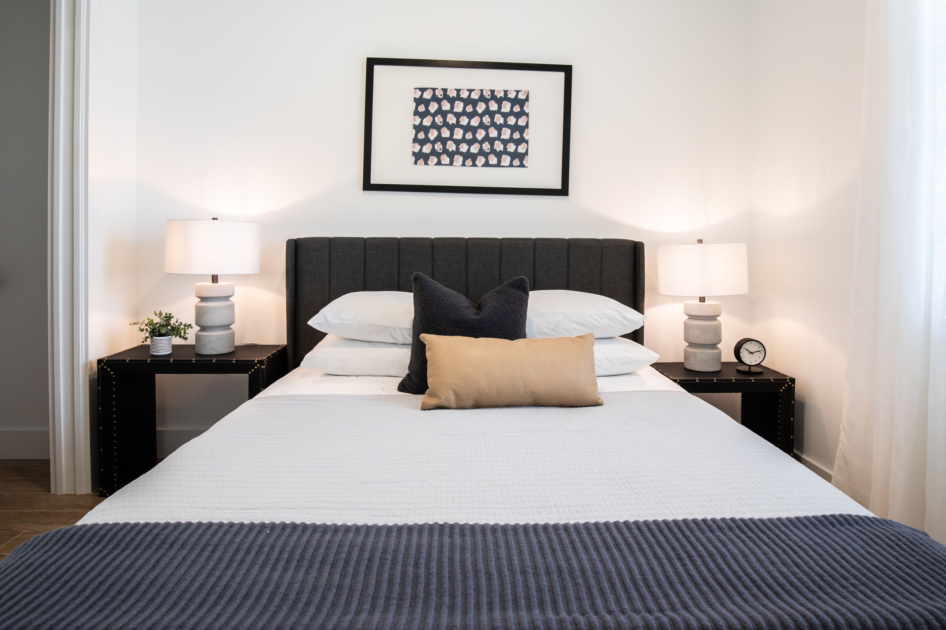 A neatly made bed with a dark headboard, two bedside tables with lamps, a framed artwork on the wall, and a window with curtains.