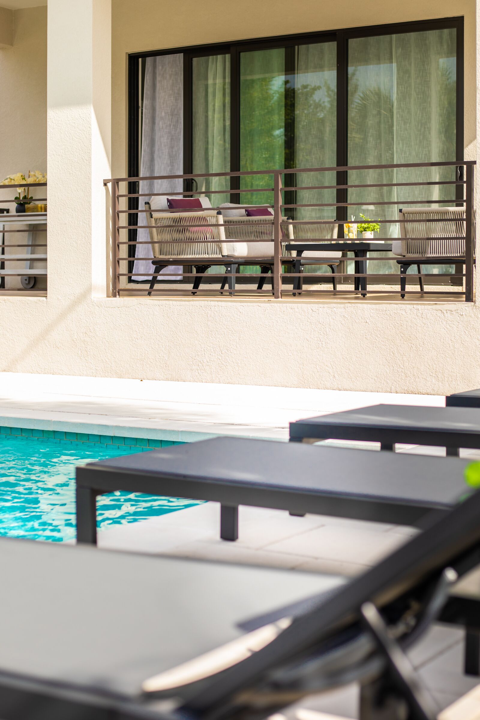 Un espace aménagé au bord de la piscine avec des chaises longues et une table, un balcon offrant une vue sur la piscine, et une fenêtre avec des rideaux.