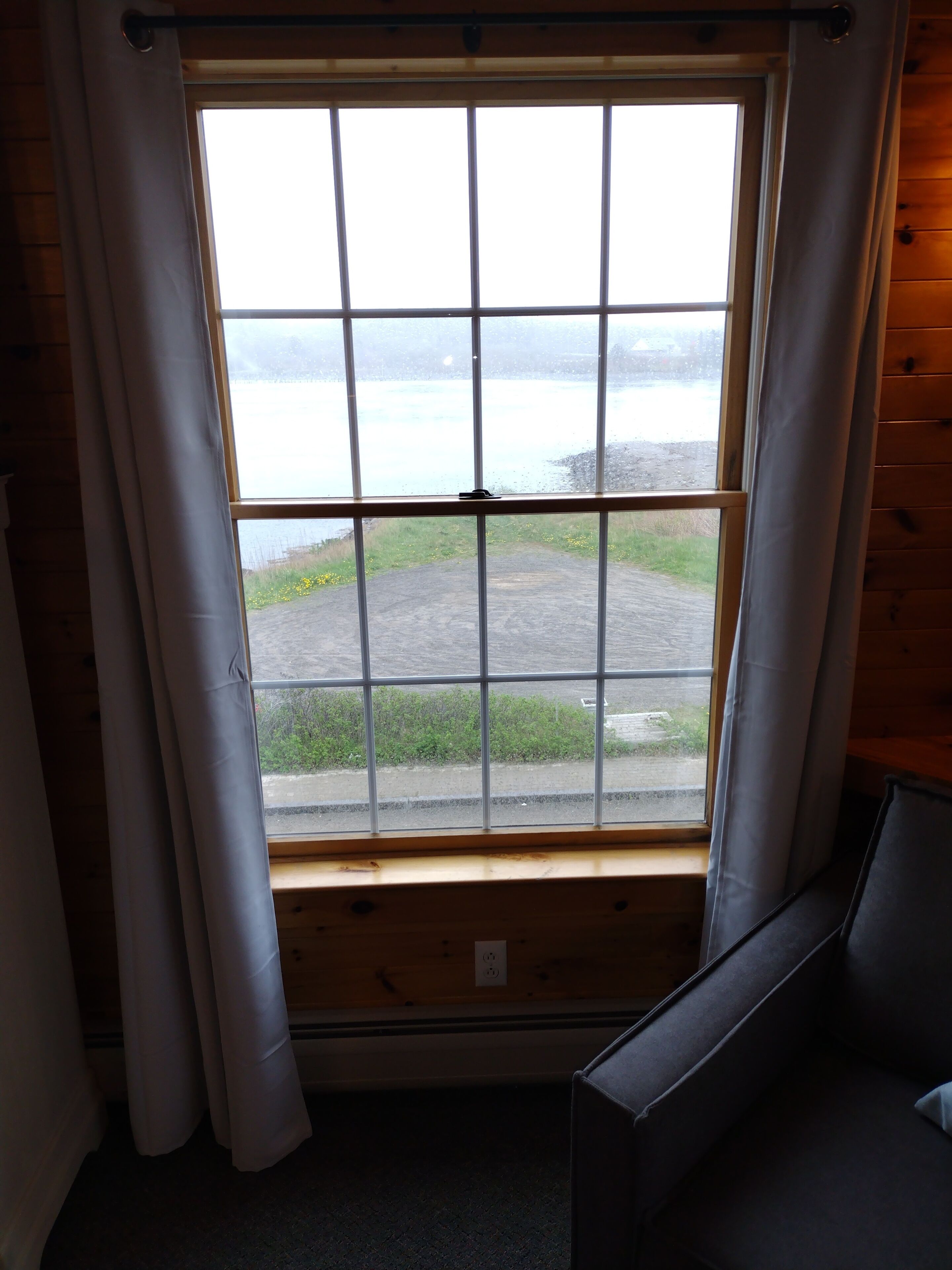 A room with a large window, wooden walls, and a view of a road and greenery outside.