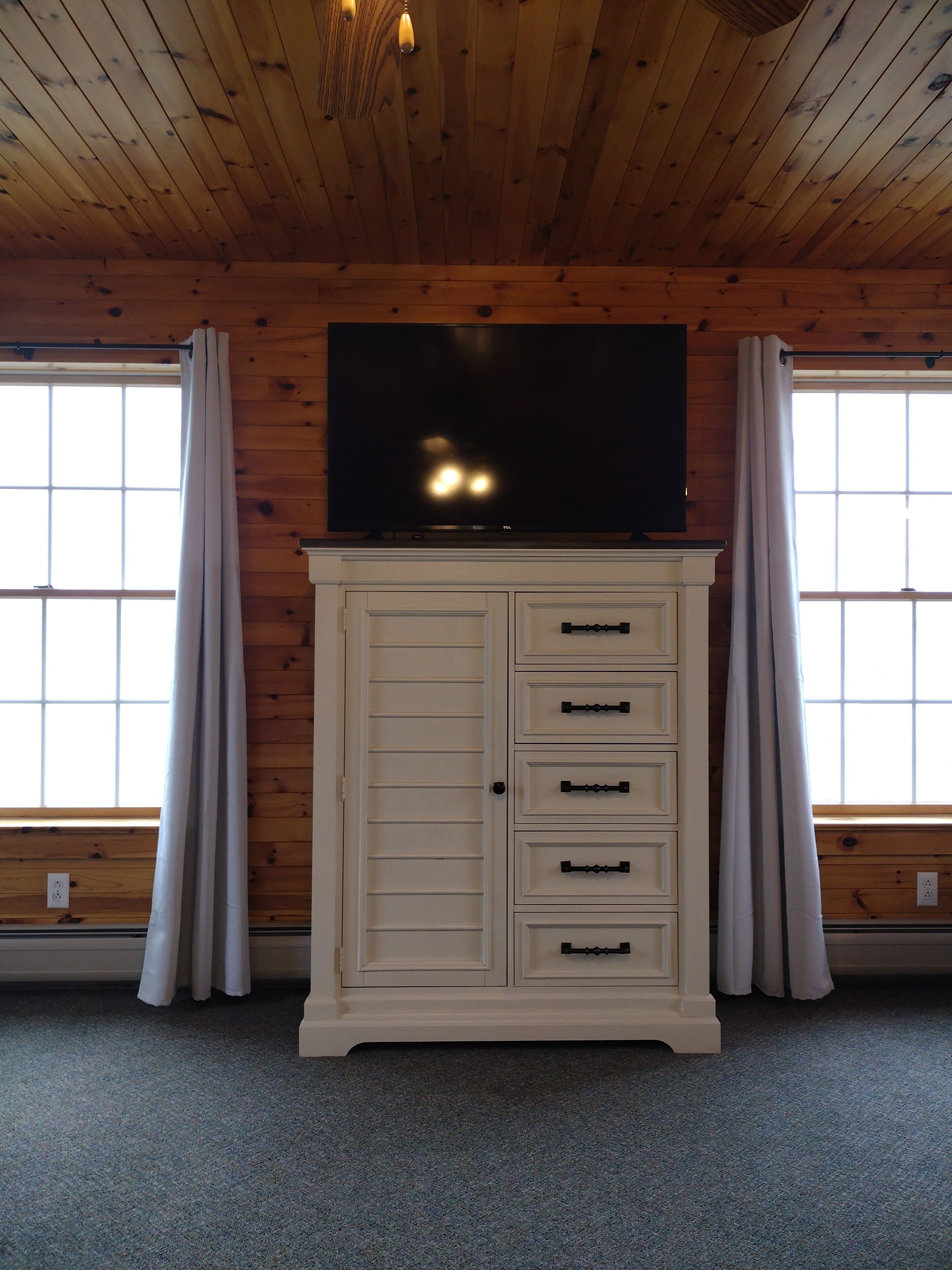 A room with a wooden ceiling, a television on a cabinet, and windows with curtains.
