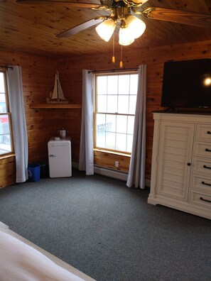 A room with wooden walls, a ceiling fan, a window with curtains, a small refrigerator, a cabinet, and a television.