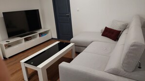 Un salon avec un téléviseur à écran plat posé sur un meuble blanc, un canapé blanc agrémenté d’un coussin rouge et une porte bleu foncé.