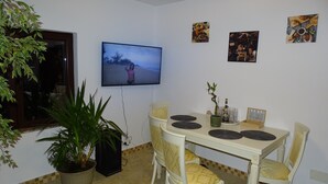 A dining area with a TV mounted on the wall, a table with chairs, a potted plant, and a small cabinet.