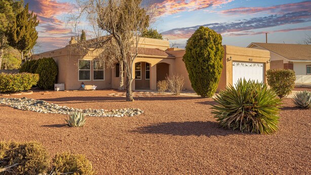 A two-story building with a beige facade, a garage, and a landscaped driveway with gravel and various plants. - Sunny, functional, Central NW --Super Clean (Albuquerque)