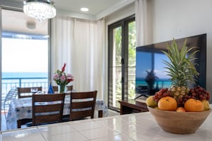 A dining area with a table set for two, a wooden bowl of fruit, and a TV on the wall.