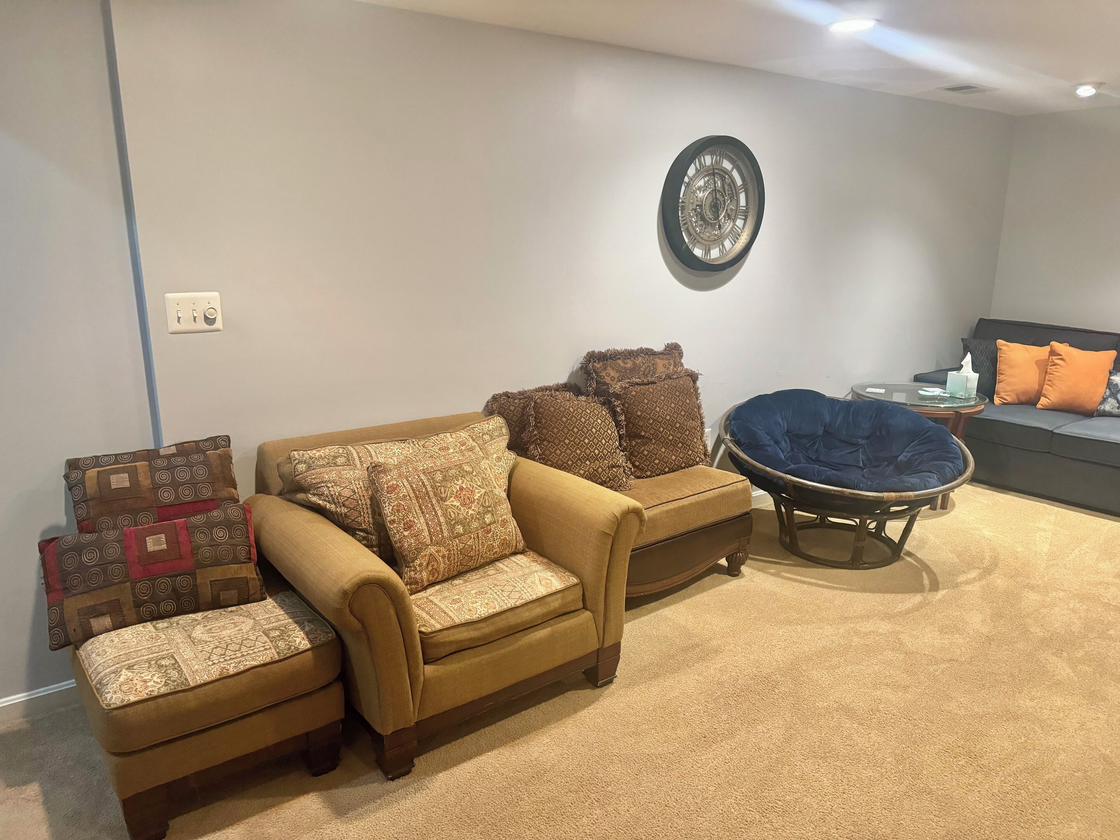 A living room with a beige sofa, patterned cushions, a blue chair, and a wall clock.