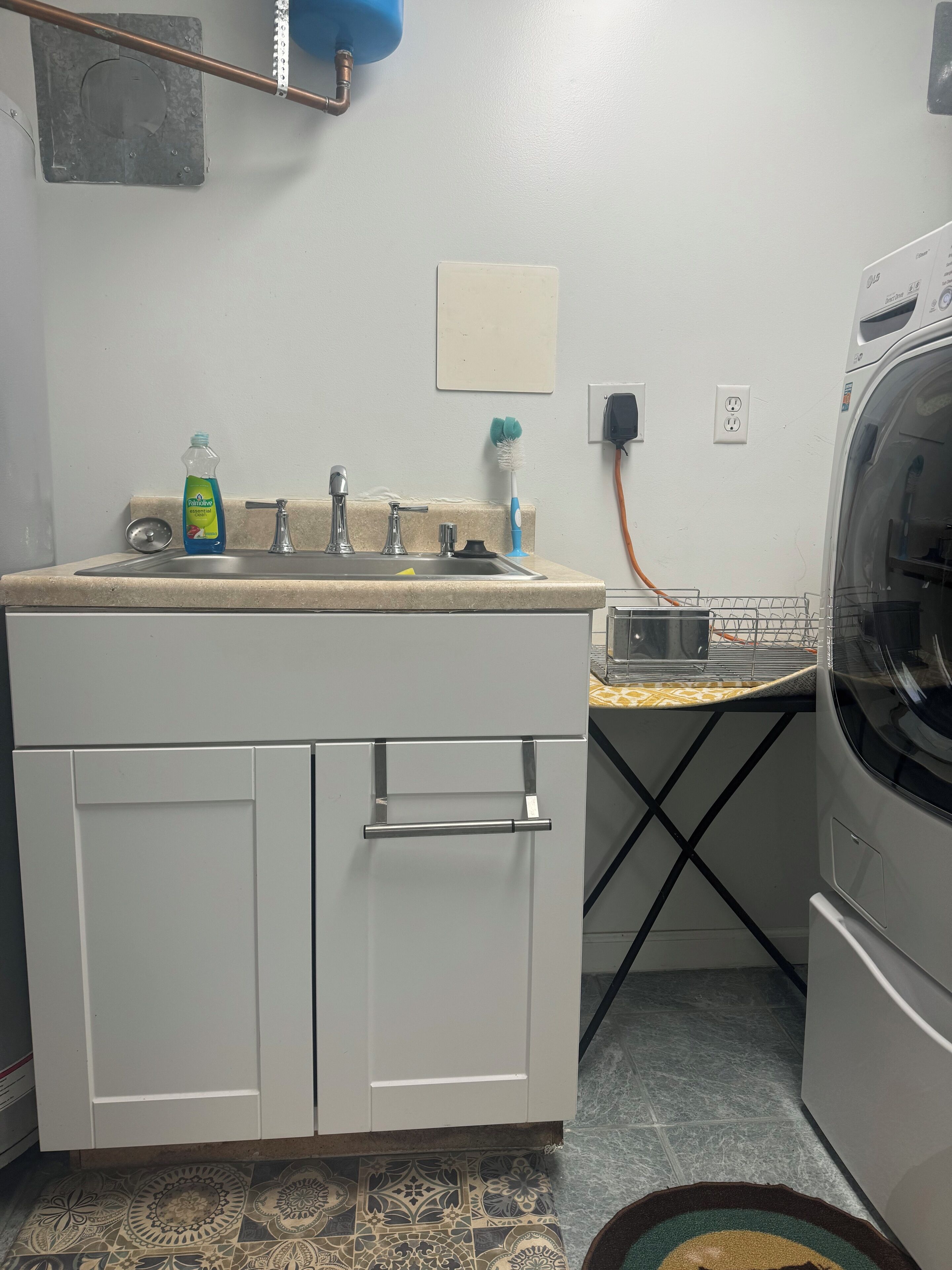 A compact kitchen with a sink, faucet, and dish soap. There is a small dish rack and a power outlet on the wall.