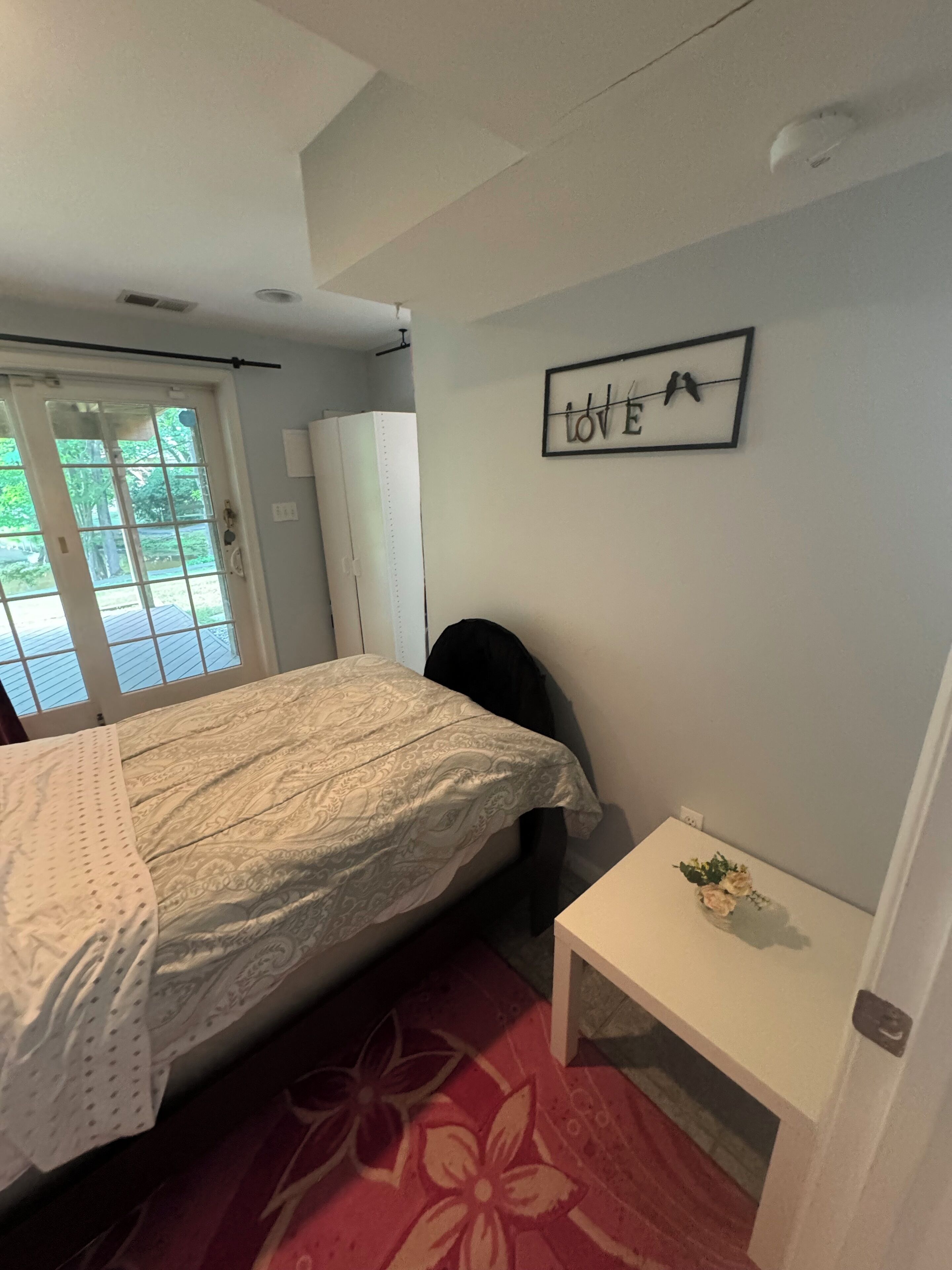 A bedroom with a bed, a desk, a chair, a small table with a flower, and a 'LOVE' sign on the wall.