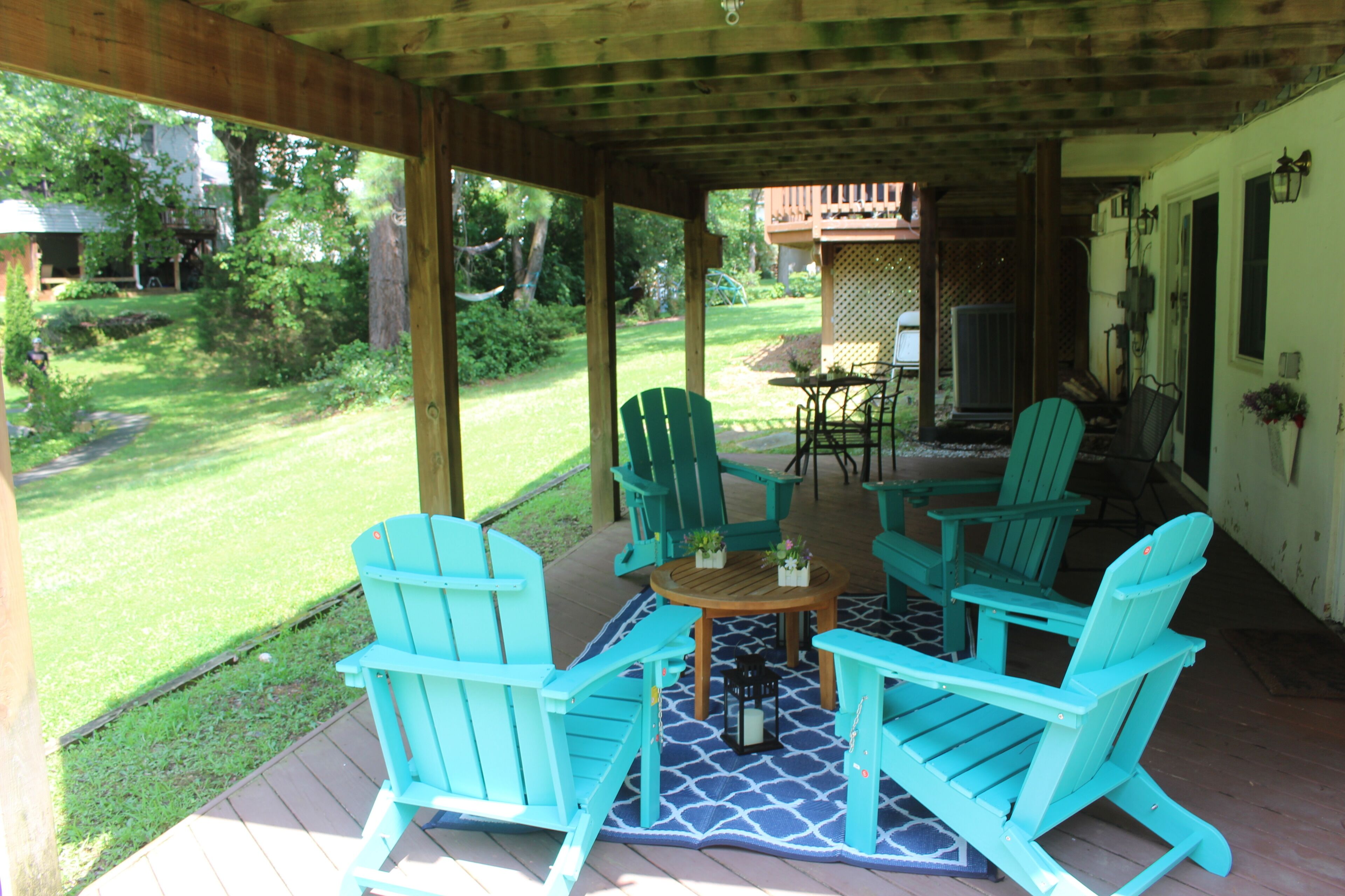A covered outdoor seating area with four turquoise Adirondack chairs and a small round table, surrounded by a wooden deck and a patterned rug.