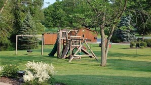A wooden play structure in a grassy yard with a small wooden building in the background.