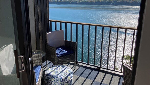A balcony with a wicker chair and ottoman, overlooking a lake with a forested hill in the background.