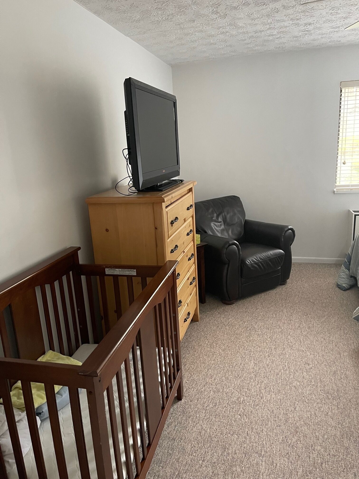 A room with a crib, a wooden dresser with drawers, a black leather chair, and a television on top of the dresser.