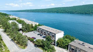 A multi-story hotel with a parking lot, situated near a large body of water with a forested hill in the background.