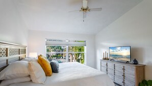 Ein Schlafzimmer mit einem großen Bett, einem Deckenventilator, einem Fernseher und Blick auf einen Pool und Palmen durch das Fenster.