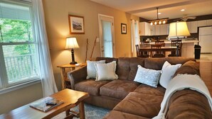 Ein Wohnzimmer mit einem braunen Ecksofa, einem Holztisch und einer Lampe. Im Hintergrund ist ein Essbereich mit Tisch und Stühlen zu sehen.