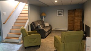Ein Wohnzimmer mit Treppe, einer Couch, zwei grünen Sesseln, einer Lampe, einem Fernseher und einem Heizgerät.