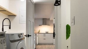 A laundry area with a washing machine and dryer, a kitchen in the background, and a hallway leading to a door. - Basement at 126 · Modern 1bedroom basement apartment newly remodeled (Kirkland)