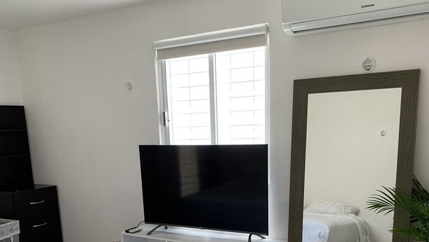 A hotel room with a bed, a TV on a stand, a window with blinds, a ceiling fan, and a large mirror. - Casa Puerta del Sol ☀️Cozy 2bedroom Wifi. AC and TV (Mérida)