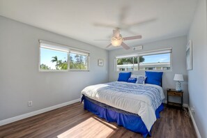 Un dormitorio con una cama, dos ventanas, un ventilador de techo y una mesita de noche.