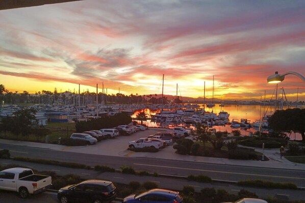 Un puerto deportivo con barcos amarrados, un estacionamiento con coches, y un atardecer sobre el agua.
