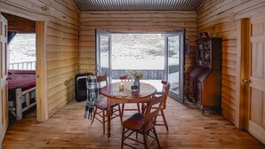 Una baita in legno con zona pranzo, tavolo e sedie, una panca e vista su un paesaggio innevato attraverso ampie finestre.
