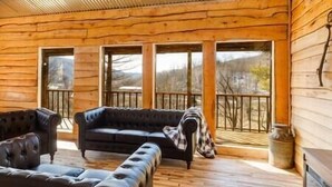 Interno di una baita di legno con un divano, poggiapiedi e una grande finestra che offre vista sul paesaggio.