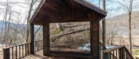 Ein hölzernes Gazebo mit geneigtem Dach und einer Holzterrasse, umgeben von Bäumen und einer Berglandschaft im Hintergrund.